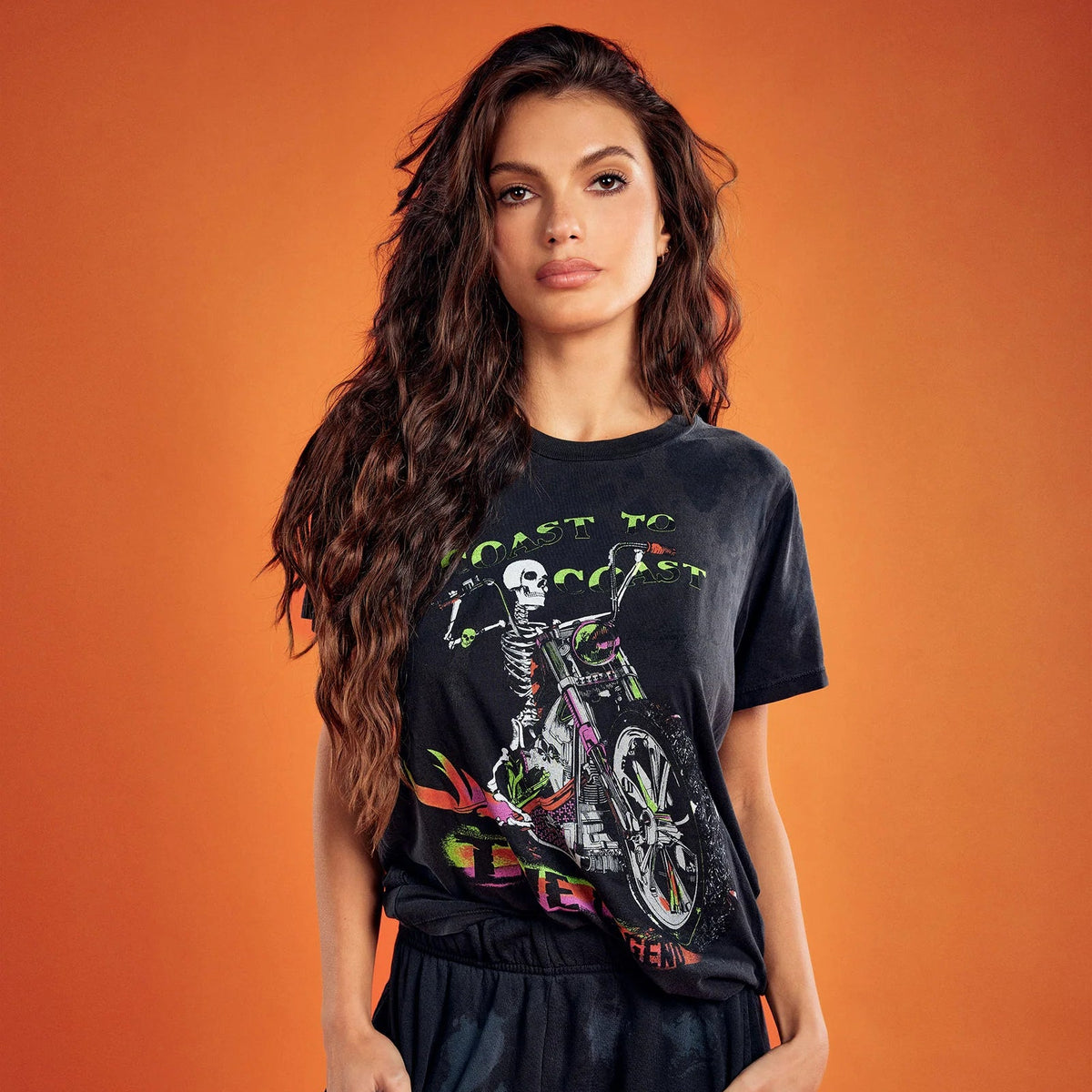 A woman modeling a black graphic t-shirt and matching black pants against a solid orange background. The t-shirt features a colorful graphic of a skeleton riding a motorcycle with green text.
