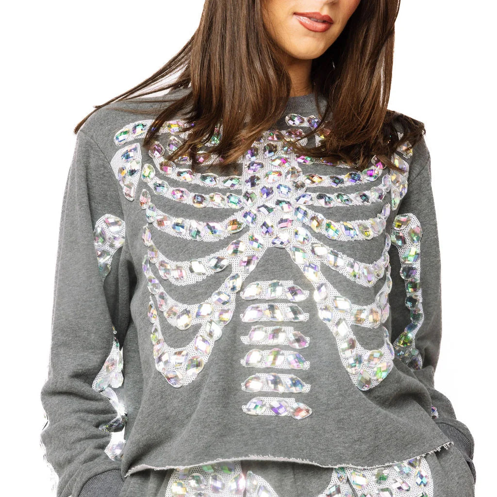 A woman wearing a gray long-sleeve sweatshirt and matching shorts set, both decorated with large white skeleton rib cage and pelvic bone patterns filled with iridescent sequins, against a plain white background.