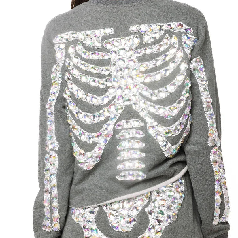 A woman viewed from the back wearing a heather gray long-sleeve sweatshirt and matching shorts, both featuring a large skeleton rib cage and hip bone graphic made from iridescent sequins.