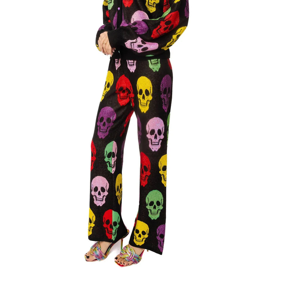 A woman modeling a black long-sleeve jumpsuit covered in a colorful skull print, paired with matching multicolor high-heeled sandals, against a plain white background.
