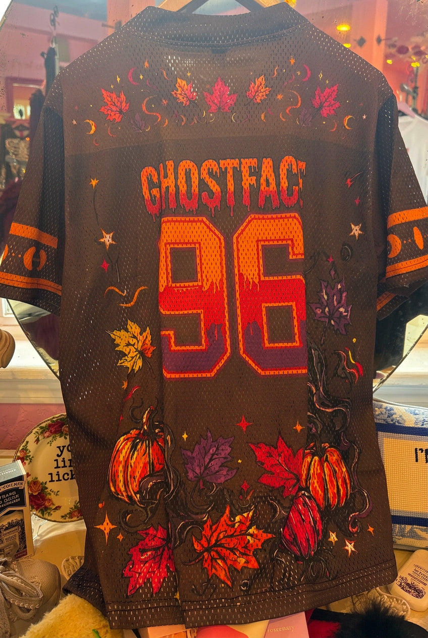 A dark brown mesh short-sleeve t-shirt with a Halloween-themed graphic print featuring the text 'GHOSTFACE 96' in orange and red, decorated with pumpkins, autumn leaves, and stars. The shirt is displayed on a wooden hanger inside a retail store window display.