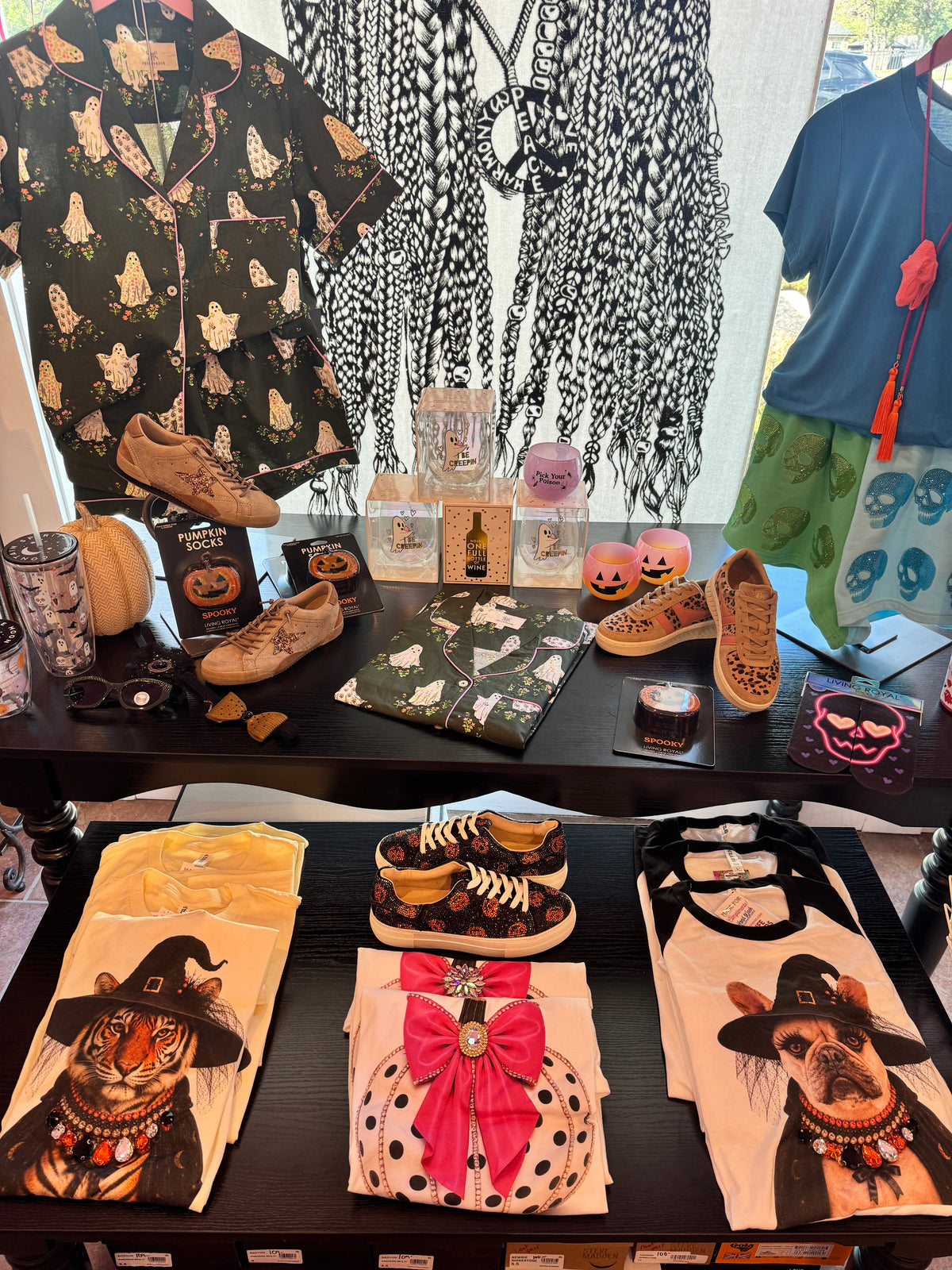A retail display table showcasing a collection of Halloween-themed apparel and accessories, including a black and white patterned pajama shirt, a blue t-shirt with green shorts, folded graphic t-shirts featuring a tiger and a bulldog in witch hats, patterned shoes, and seasonal decor.