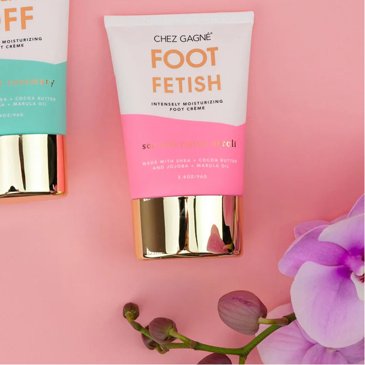 Two tubes of Chez Gagnée foot cream, one labeled 'Walk It Off' in mint green and one labeled 'Foot Fetish' in pink, arranged on a pink background with purple orchid flowers.