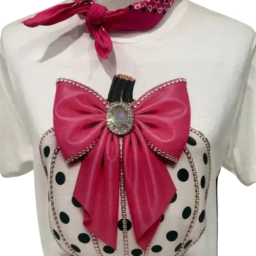 A white short-sleeve t-shirt displayed on a mannequin torso, featuring a large graphic of a pink velvet-texture bow and ribbon tied to a cream pumpkin with black polka dots and silver rhinestone trim. A matching pink neck scarf is tied at the collar, all set against a solid black background.