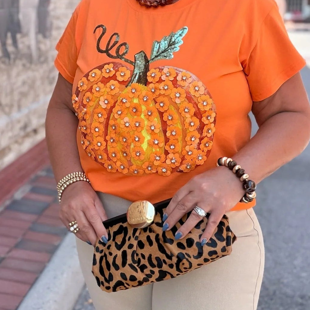 A woman wearing a bright orange graphic t-shirt with a large embellished pumpkin design and the word 'We', paired with beige pants and holding a leopard-print clutch, standing outdoors next to a brick wall.