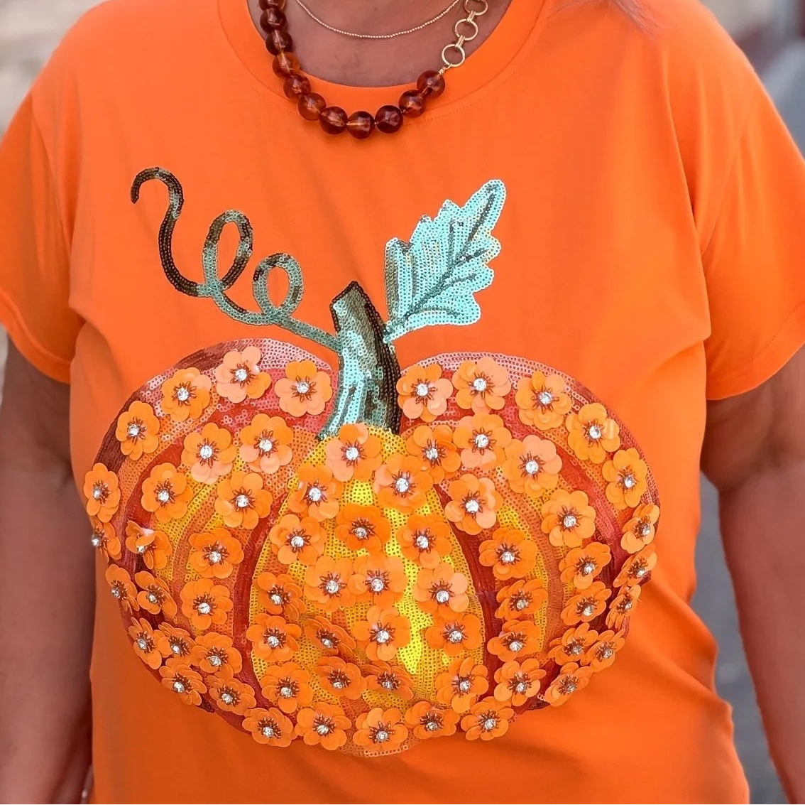 A woman wearing a bright orange t-shirt with a large 3D pumpkin applique on the front, standing outdoors against a stone wall.